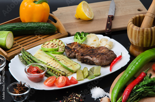 Grilled beef grilled steak with asparagus, zucchini, lobi, broccoli, tomato, chili and red sauce on a white plate, on a black glossy background