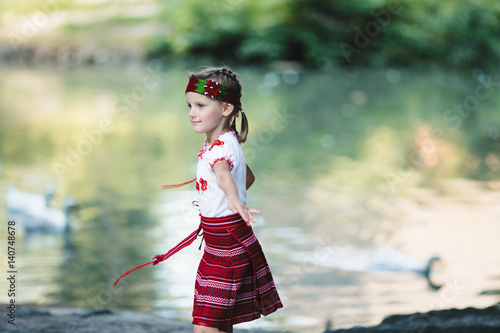 Fototapeta Naklejka Na Ścianę i Meble -  Charming little girl in embroidered clothes whirls before the lake