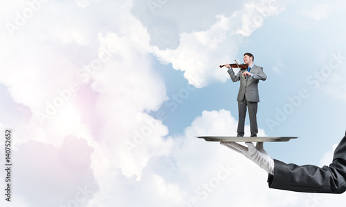 Businessman on metal tray playing violin against blue sky background © adam121