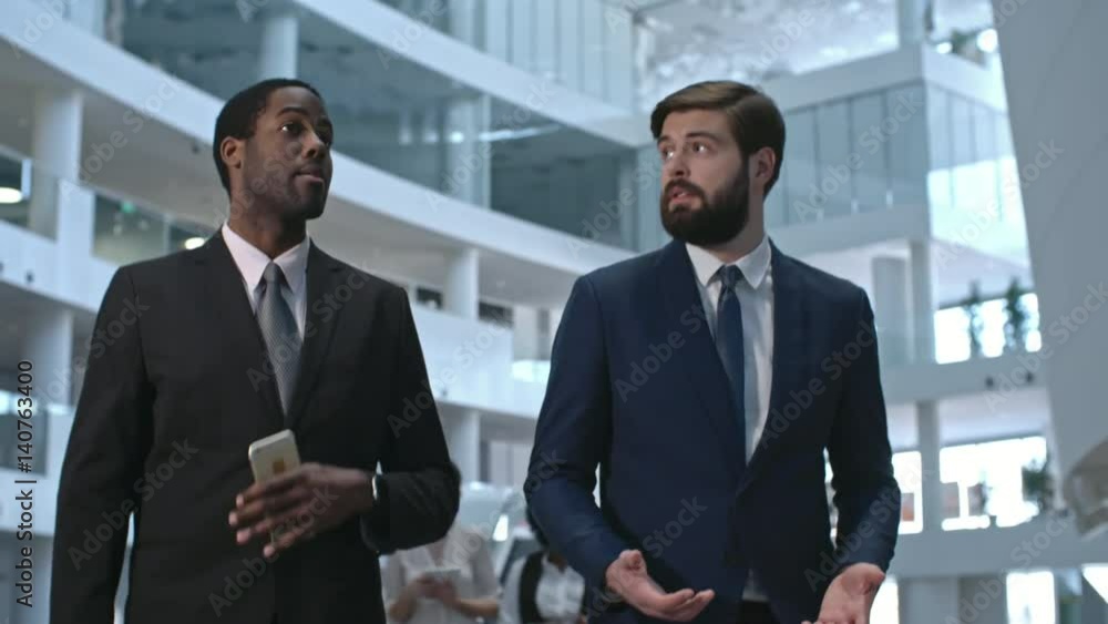 Dolly shot of African businessman in suit walking through lobby of modern business center and discussing work with gesturing bearded colleague Dolly shot of African businessman in suit walking through lobby of modern business center and discussing work with gesturing bearded colleague