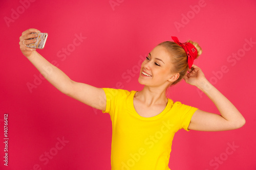 Beautiful young blond teenage woman in yellow t shirt taking selfie over pink background.