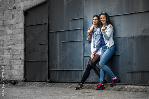 Portrait of two young and attractive women standing next to the wall, looking at camera. Barcelona, Spain. One girl is showing the middle finger at camera.