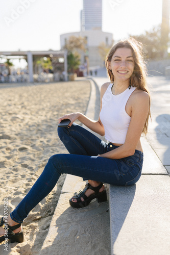 Spain, Barcelona, portrait of smiling young woman sitting on beach promenade at sunset