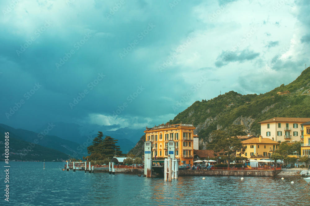 Beautiful cityscape of coastline of italian Varenna city with Como lake, building, big trees, green mountain and dramatic sky