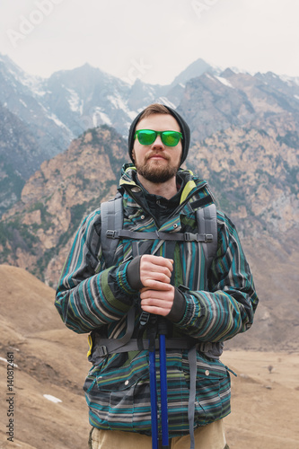 A guy with a beard and wearing sunglasses in a membrane jacket, hat, with a backpack and sticks for Nordic walking, a traveler standing and looking at the mountains