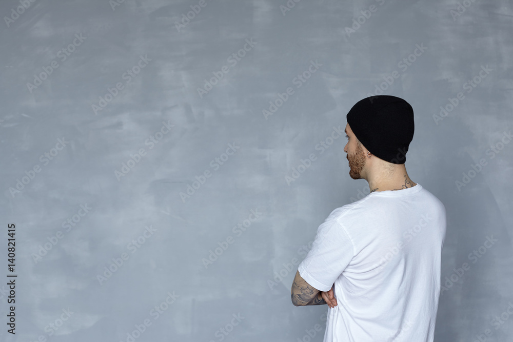 Fototapeta premium Back view on man in white t-shirt and black hat look to grey wall. Brutal bearded hipster on neutral background. Person watch on big blank space.