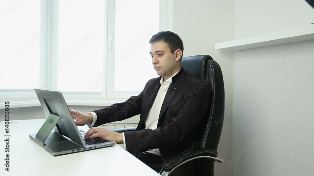 Businessman sitting in an office chair at the table and typing on the silver laptop. Businessman sitting in an office chair at the table and typing on the silver laptop.
