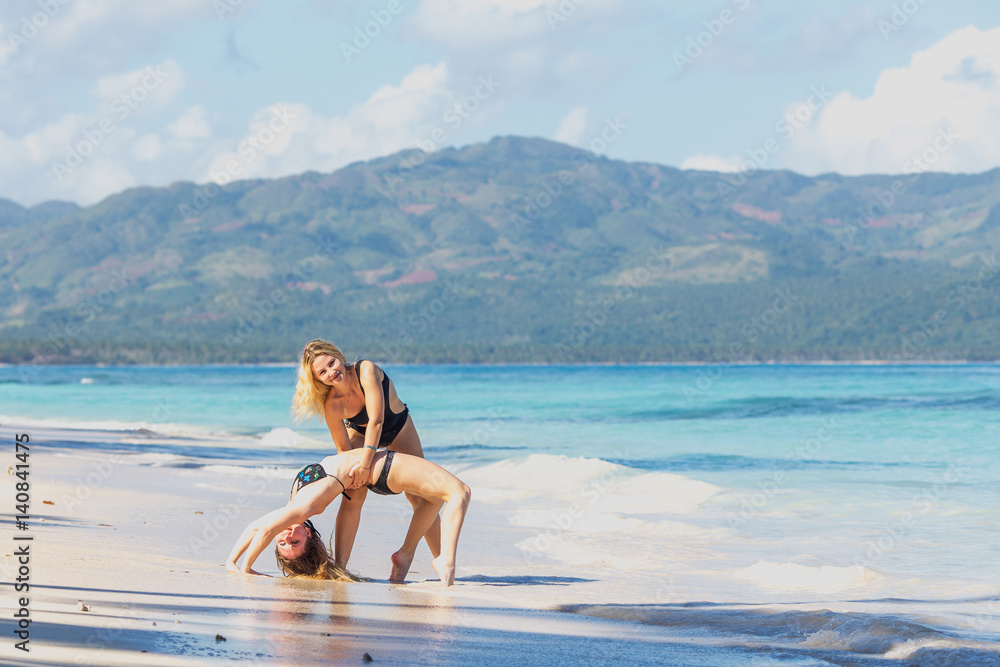 two pretty young girls in bikini exercising bridge pose yoga on the beach