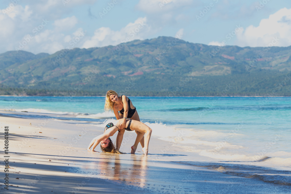 two pretty young girls in bikini exercising bridge pose yoga on the beach