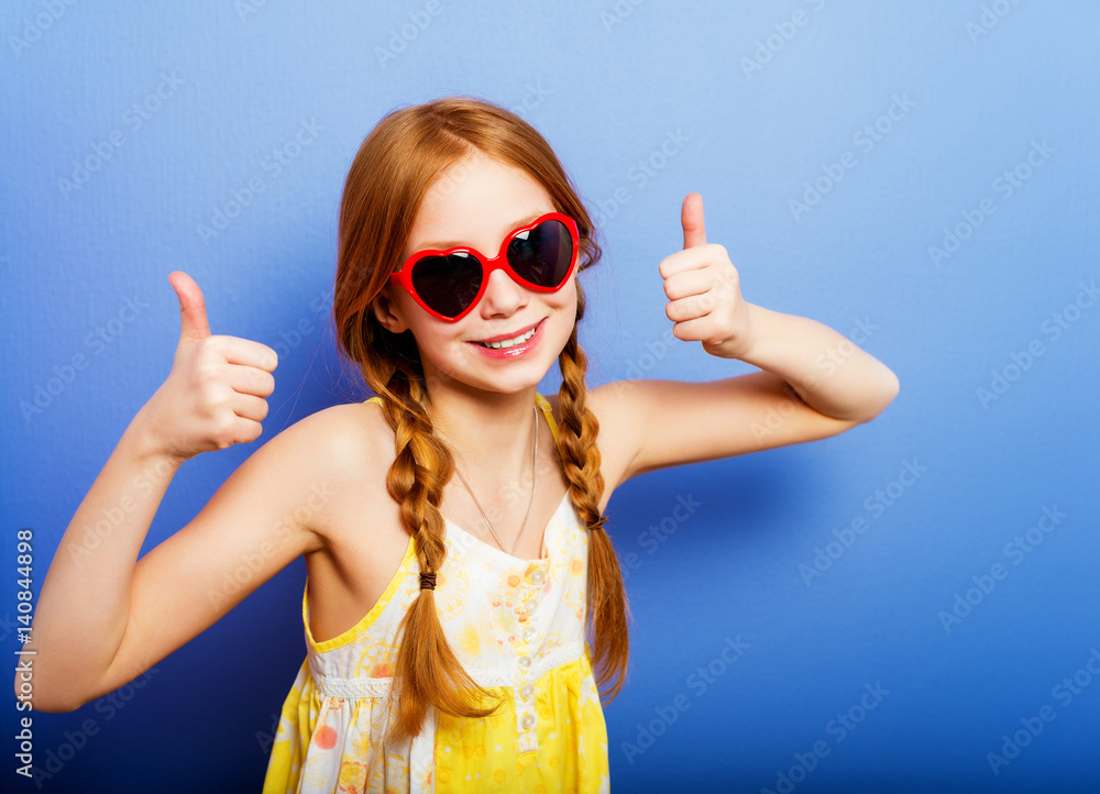 Studio shot of young preteen 9-10 year old redhead girl wearing heart shape sunglasses, standing against blue purple background, big thumbs up