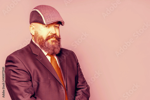 Adult bearded man in a suit and cap on a pink background