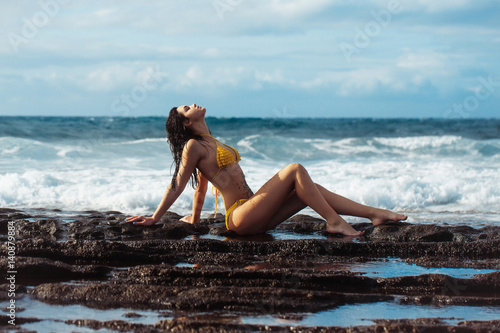 Pretty girl in sexy yellow swimsuit sitting on rocky beach