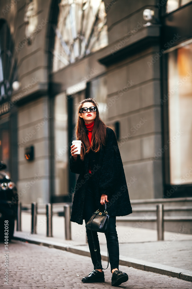 young stylish girl, passing by the Windows.Wearing fashionable glasses and a black coat.Keeps coffee.