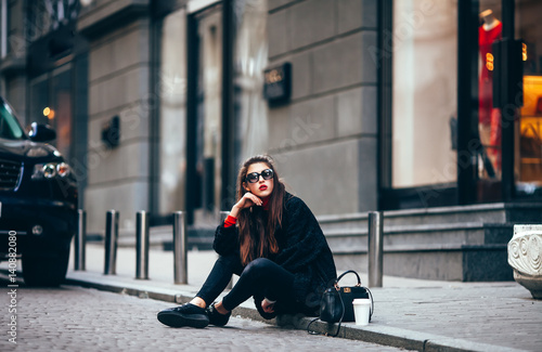 young stylish girl, passing by the Windows.Wearing fashionable glasses and a black coat.Keeps coffee. looks at camera