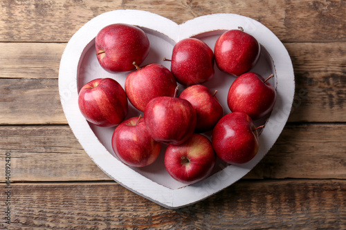 Plate in shape of heart with fresh red apples on wooden background