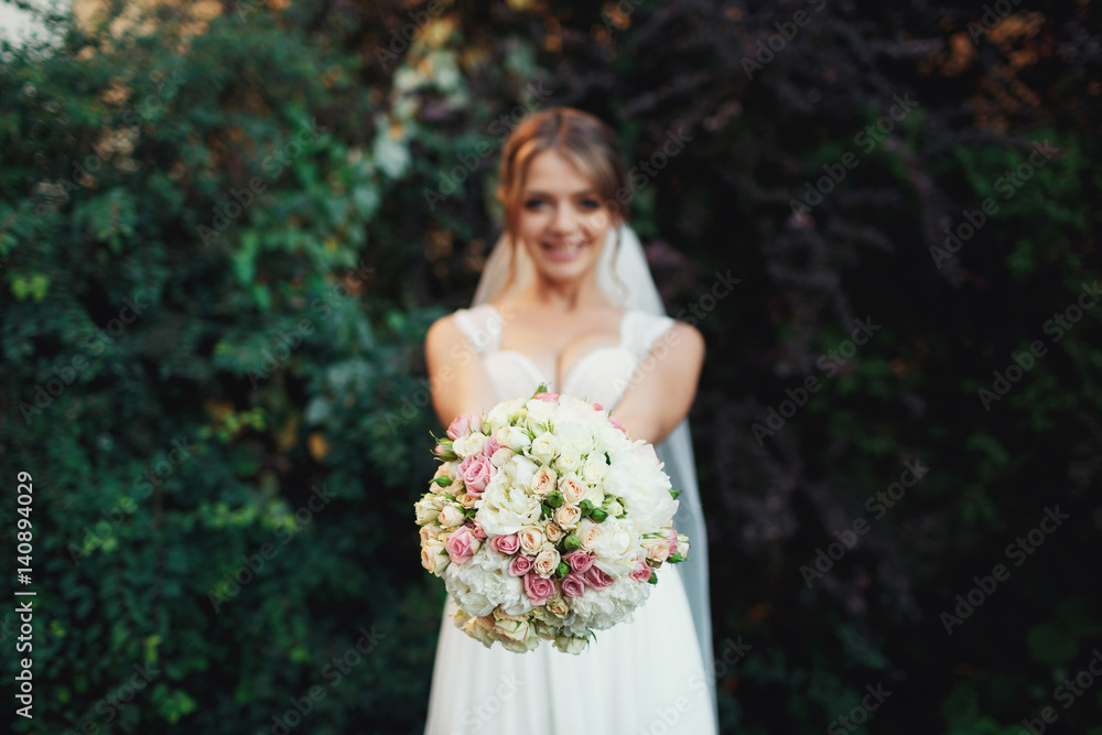 Bride reaches out her hands with pink wedding bouquet standing before green bushes Bride reaches out her hands with pink wedding bouquet standing before green bushes