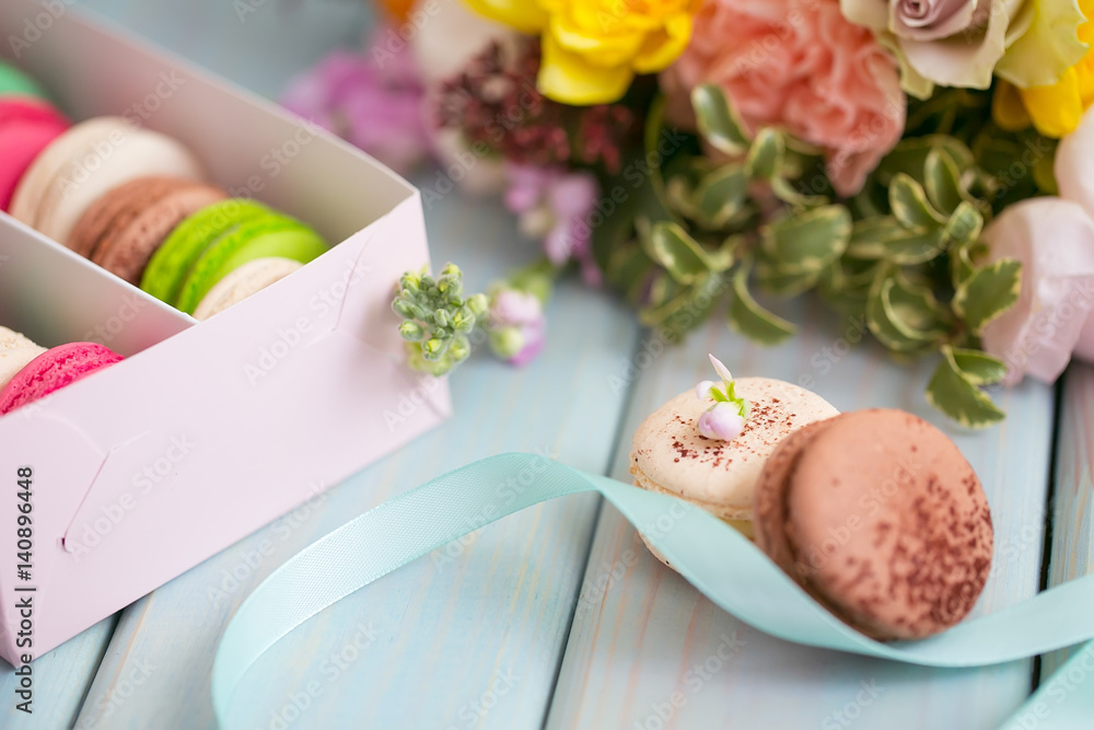Box with colored macaroon cake, a bouquet of flowers on a blue wooden background.