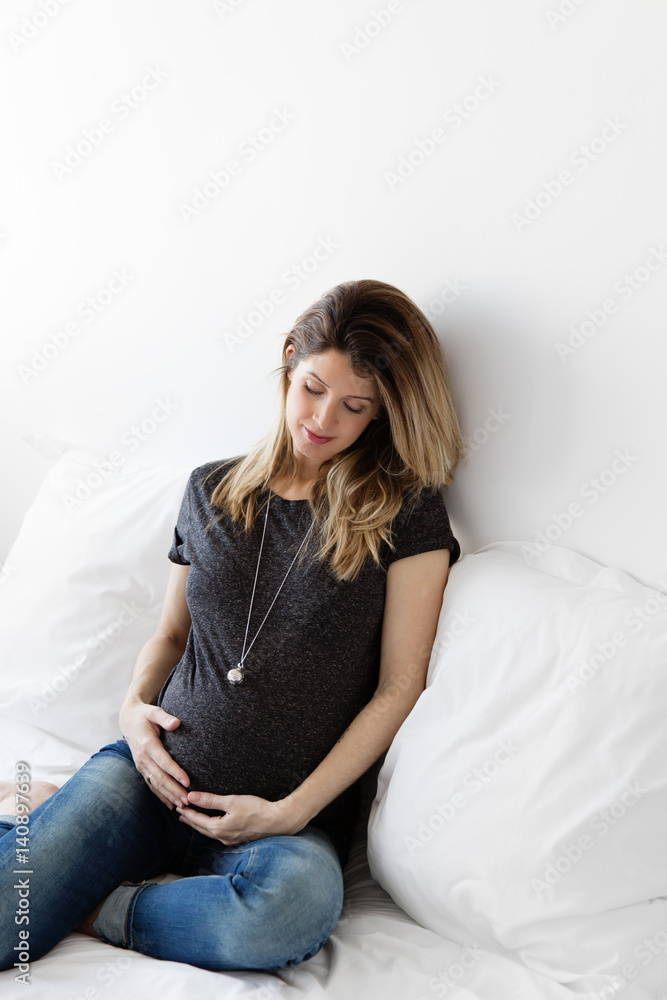 pregnant woman sitting on a white bed with a bola necklace looking at her stomach pregnant woman sitting on a white bed with a bola necklace looking at her stomach