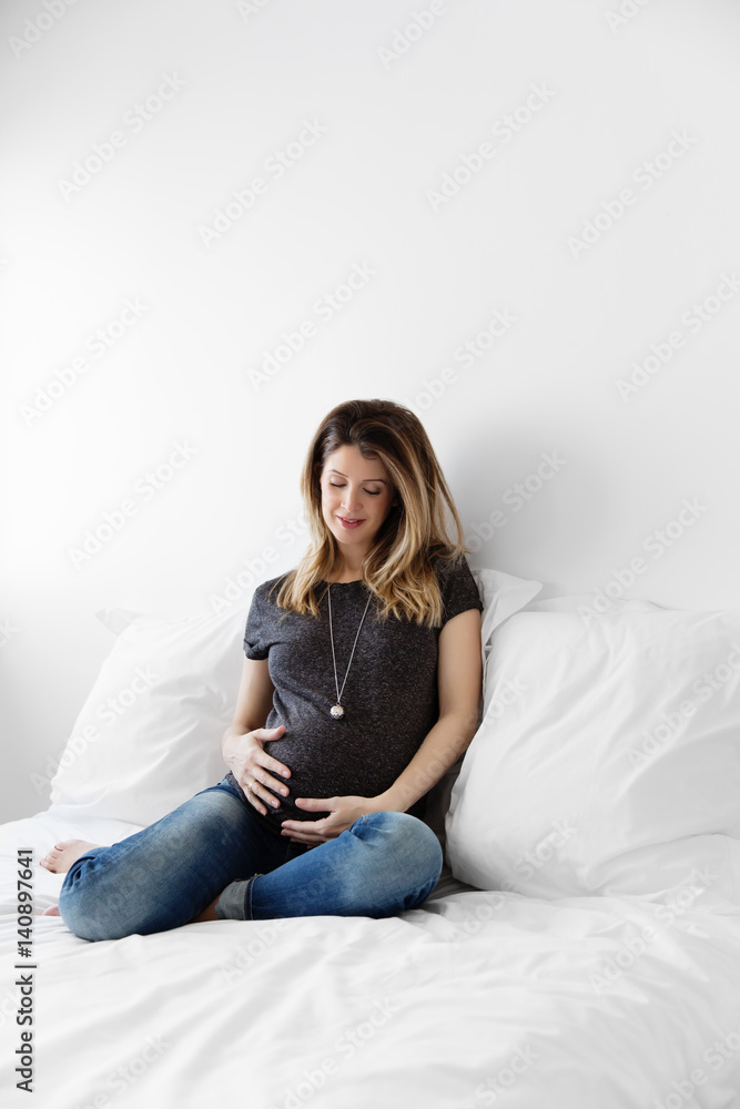 pregnant woman sitting on a white bed with a bola necklace looking at her belly pregnant woman sitting on a white bed with a bola necklace looking at her belly