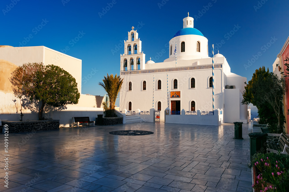Picturesque square and white church of Panagia Platsani with blue dome in Oia or Ia on the island Santorini in the morning, Greece