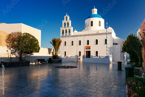 Picturesque square and white church of Panagia Platsani with blue dome in Oia or Ia on the island Santorini in the morning, Greece