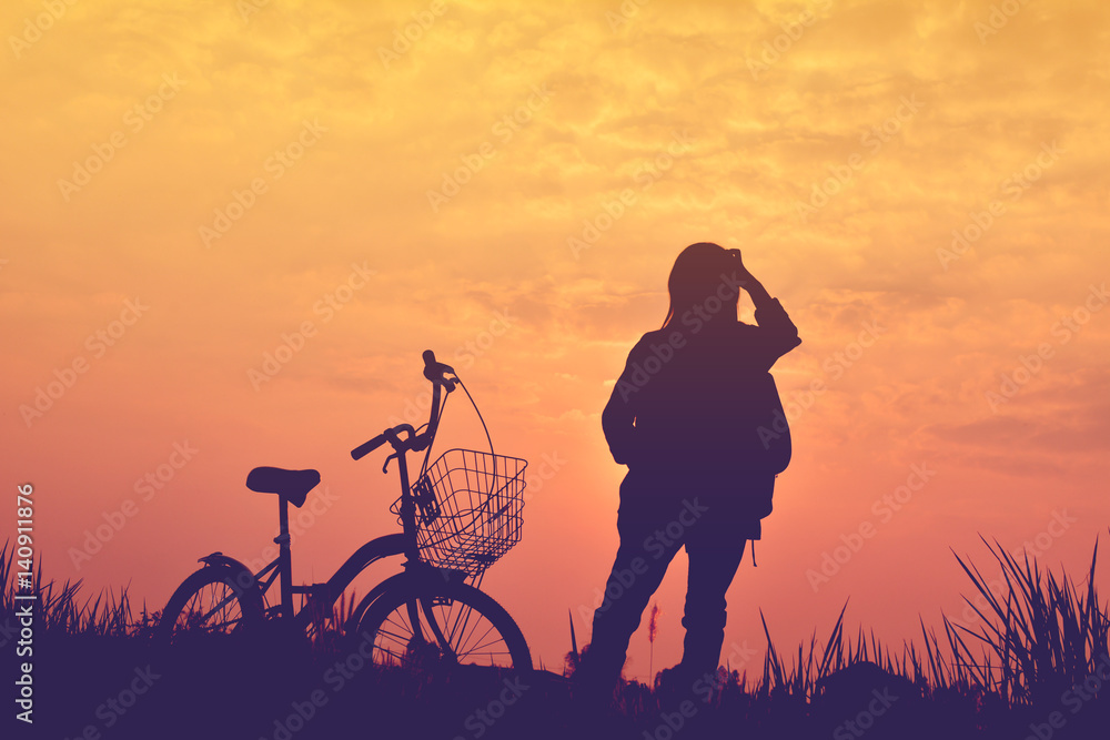 Fototapeta premium Silhouette of girl with bicycle on grass field at the sky sunset, color of vintage tone and soft focus concept journey