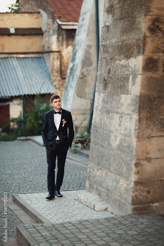 Tall groom in black tuxedo walks along the stone wall