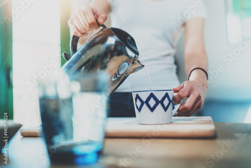 Young woman using kettle for make tea or black coffee on kitchen at room interior background.Women's hands pour water from a teapot into a cup. Blurred, flares effect. photo