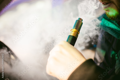 young vaper man with beard vaping mechanical mod. Guy smokes an electronic cigarette by blowing a smoke vapor. Holds in hand on colorful background