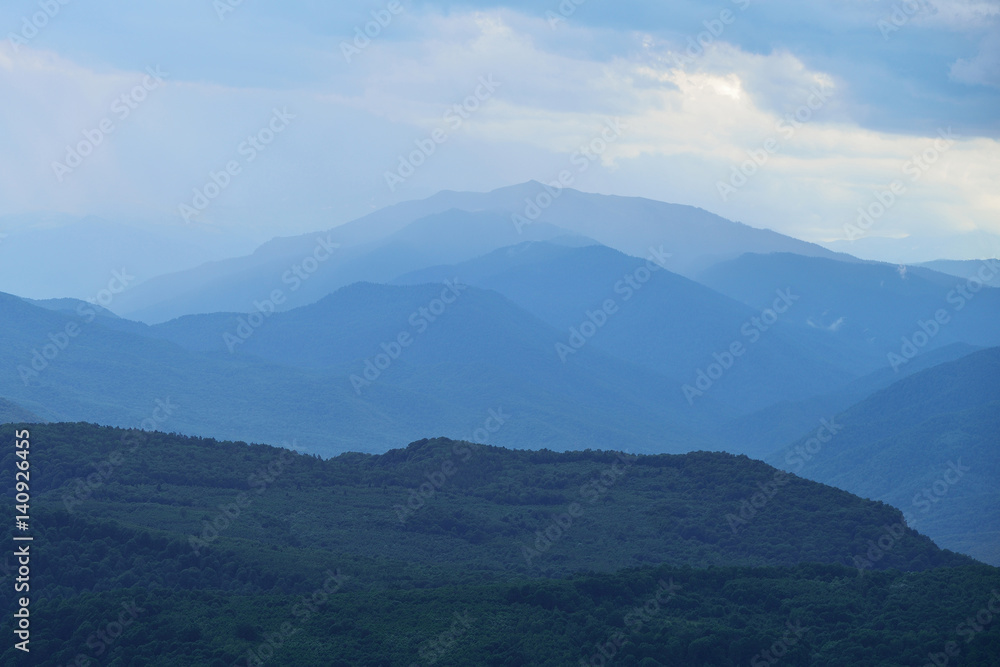 Blue nature background with mountain silhouettes and a pine tree forest. Smooth gradient from dark to light blue caused by the long-focus lens and twilight.