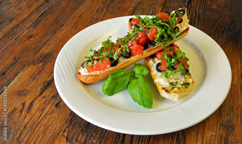 Preparing delicious Italian tomato bruschetta with chopped vegetables, olives, cheese and oil on grilled or toasted crusty baguette sprinkled with seasoning and spices on a plate on wooden background