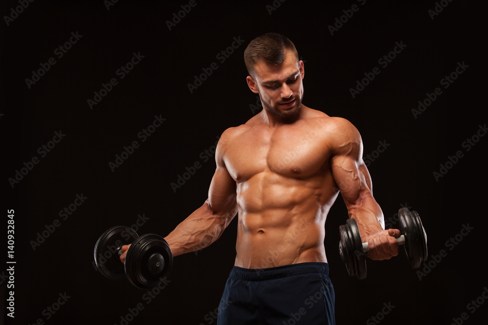 Handsome athletic man in gym is pumping up muscles with dumbbells in a gym. Fitness muscular body isolated on dark background.