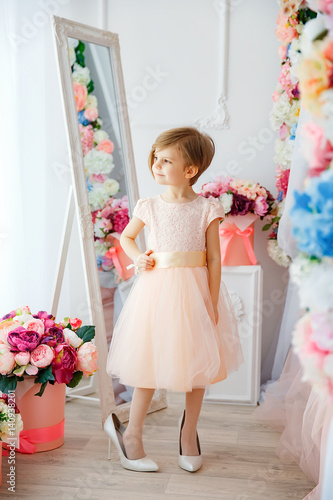 Little adorable girl in dress and female high heels posing and looking away in room filled with flowers. © zamuruev