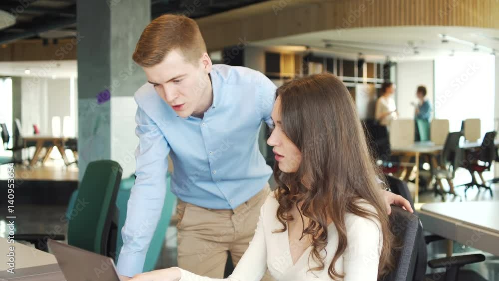 young working man stay near sitting working young woman with long dark hair and solve business questions using laptop computer, discussing and speaking in the office young working man stay near sitting working young woman with long dark hair and solve business questions using laptop computer, discussing and speaking in the office