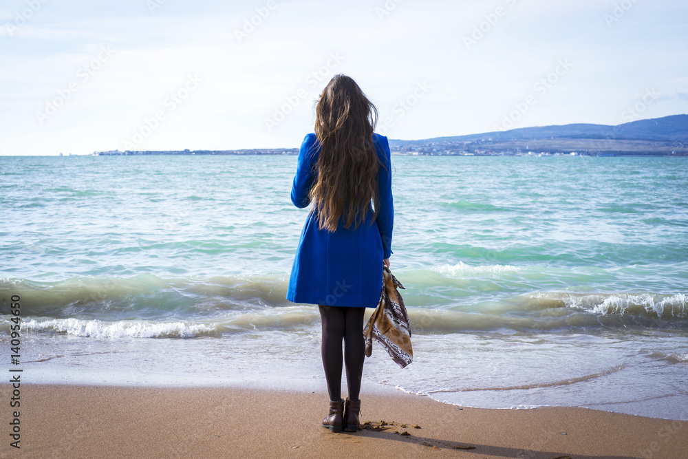 Стоящая спиной девушка на берегу моря с длинными волосами в синем