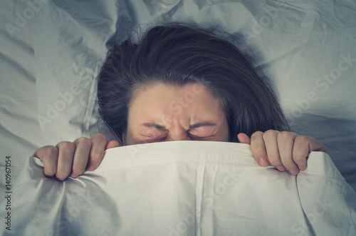 Woman with stressed face expression eyes closed hiding behind sheet in bed photo