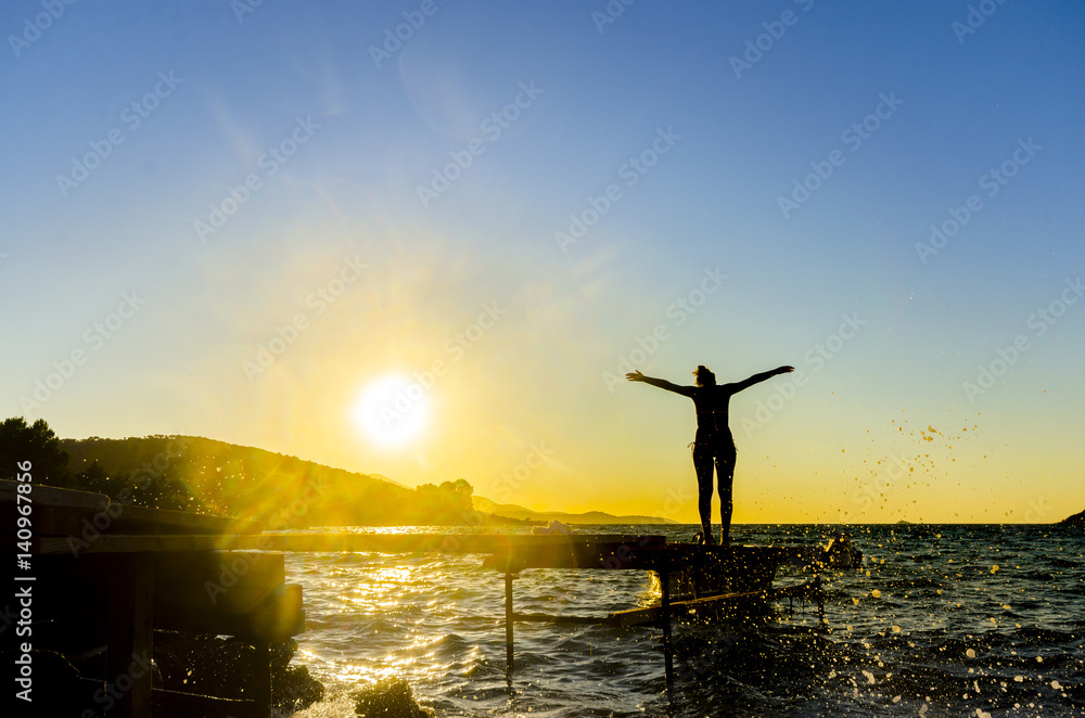Portrait of healthy young hispanic woman standing on the beach pier with her hands outstretched against sunset. Wave water splashes from rocky coas are visible in foreground