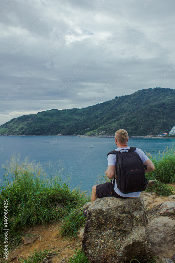 Traveller sitting near the ocean. Back view of a traveller sitting and looking at the seascape. Vertical outdoors shot. Traveller sitting near the ocean. Back view of a traveller sitting and looking at the seascape. Vertical outdoors shot.