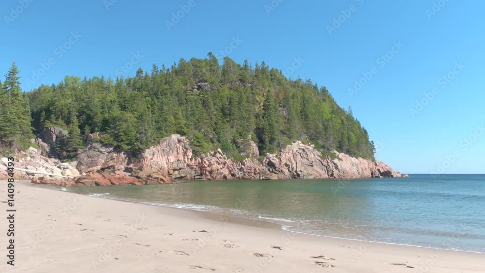 CLOSE UP: Small ocean waves washing pristine sandy northern beach in Canada. Dangerous rocky cliffs covered with early spring pine, spruce and fir foliage in natural forest rising above the calm sea CLOSE UP: Small ocean waves washing pristine sandy northern beach in Canada. Dangerous rocky cliffs covered with early spring pine, spruce and fir foliage in natural forest rising above the calm sea