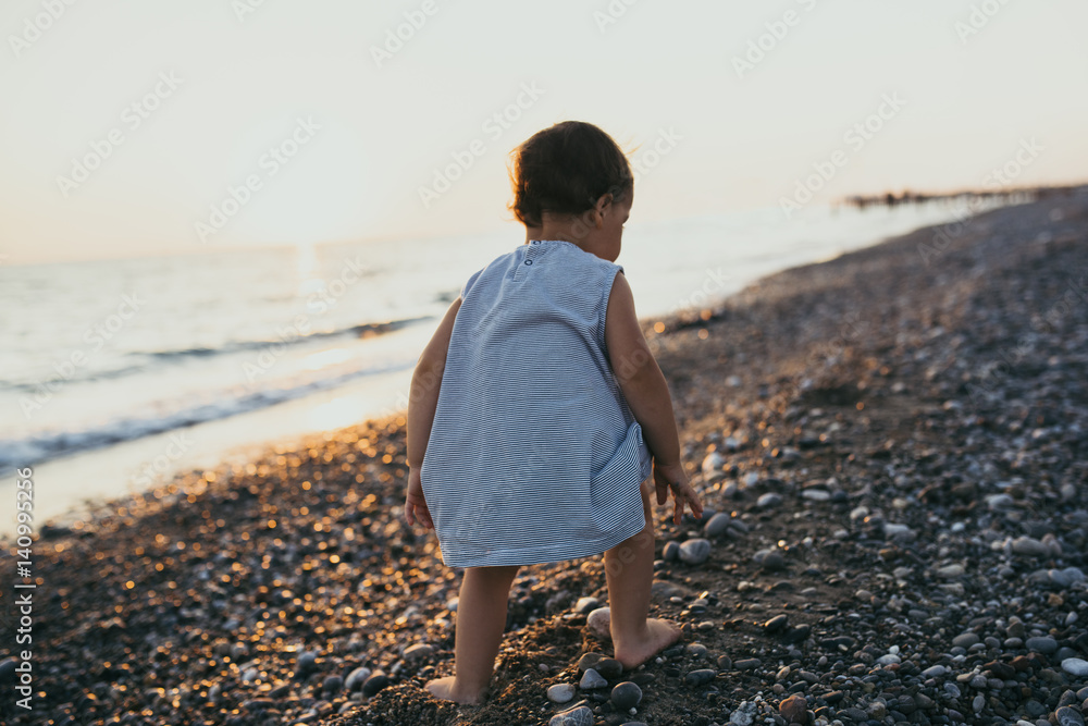 Back view cute girl toddler play at the sea beach sunset background. Happy familly summer vacation. Travel. Caucasian happy baby sit play game activity at ocean sunset Childhood lifestyle. Silhouette.
