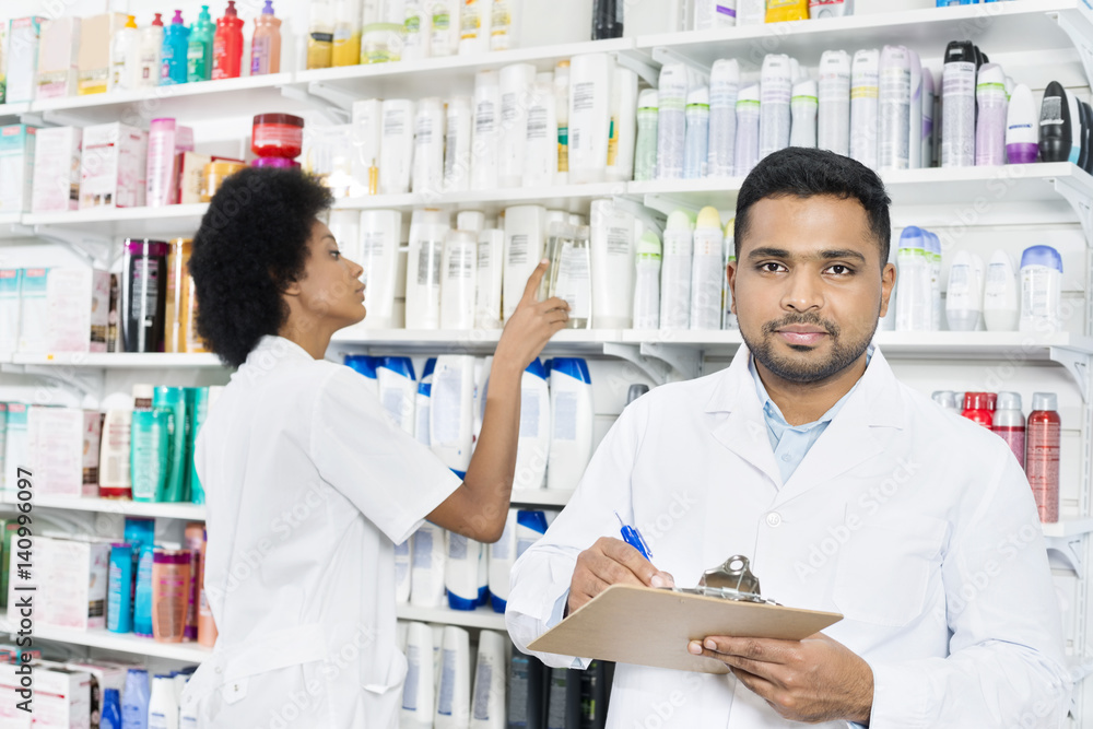 Male Pharmacist Holding Clipboard While Colleague Arranging Stoc