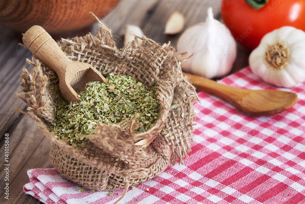 Dried oregano spice in burlap bag with wooden scoop, garlic and tomato on table. Concept image for cooking with oregano