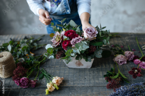 female  in gray blouse and jeans make a bouquet over gray background, putting roses in vase, flowers and vase on wood table, workplace