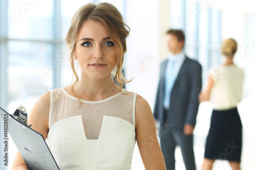 Blonde woman with touchpad computer looking at camera and smiling while business people are shaking hands over office background