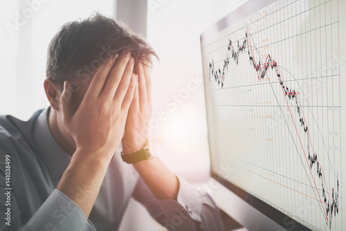 Frustrated stressed shocked business man with financial market chart graphic going down on grey office wall background. Poor economy concept. Face expression, emotion, reaction photo