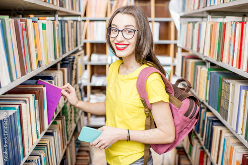 Young happy and enthusiastic female student searching books on the shelves at the library