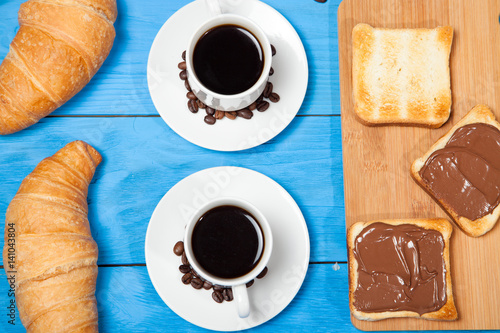 Two cups with coffee, croissant and toast with chocolate on a blue wooden table