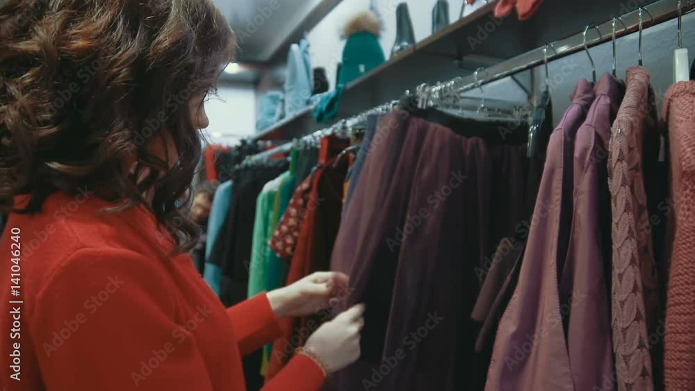 A young brunette woman chooses a suit in a ready-to-wear shop, sorting out things on a hanger. A young brunette woman chooses a suit in a ready-to-wear shop, sorting out things on a hanger.