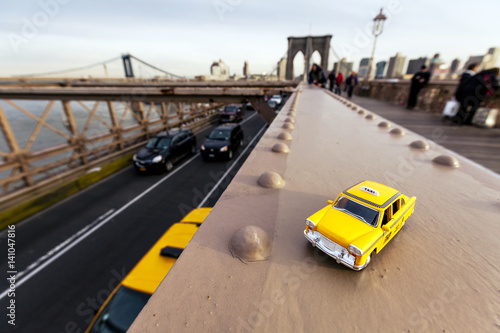 Model of a classic yellow taxi on a steel beam on Brooklyn Bridge, with a real taxi passing on the lower level asphalt road.