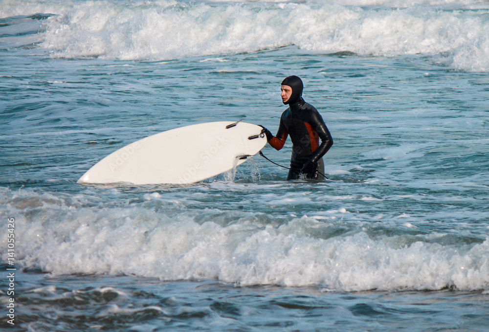Naklejka premium Surfer walks from water with surfboard wearing a waterproof suit.